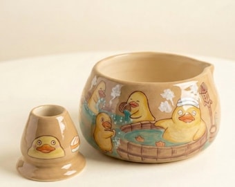 May include: A beige ceramic bowl and a matching small stand, both featuring illustrations of yellow ducks. The bowl depicts ducks bathing in a wooden tub with steam, while the stand features a single duck face and a small white food item.