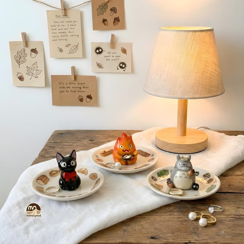 May include: A wooden table displays a small lamp with a beige shade, three decorative ceramic dishes with character figurines, and several rings. The dishes feature illustrations and hold jewelry. Notes are pinned above.