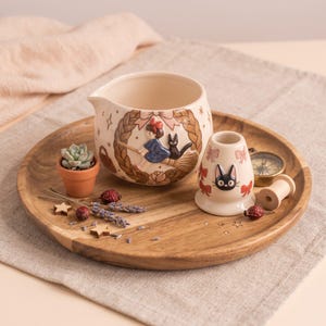 Może przedstawiać: Ceramiczny dzbanek na śmietankę oraz mały, pasujący świecznik z motywem dziewczynki na miotle i czarnego kota. Dzbanek posiada wzór plecionego wieńca, natomiast świecznik ozdobiony jest kocim pyszczkiem oraz czerwonymi kokardkami.