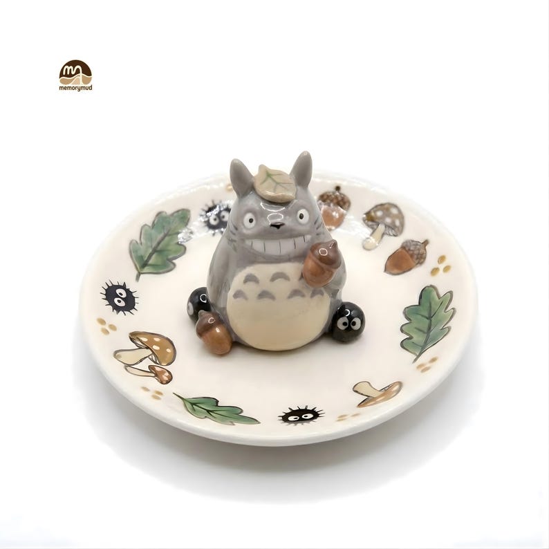 May include: A ceramic decorative plate featuring a gray Totoro figure holding an acorn. The plate is decorated with painted leaves, mushrooms, and other forest-themed elements. The plate is white with a slightly raised edge.