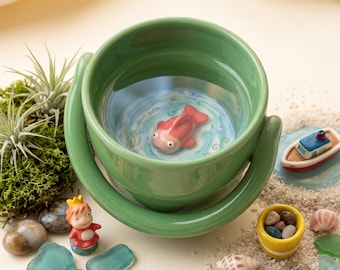 May include: A sage green ceramic bowl with a handle, featuring a three-dimensional pink fish figurine at the bottom. The interior base is painted with blue water ripples. The bowl is surrounded by small decorative items including a tiny boat and shells.