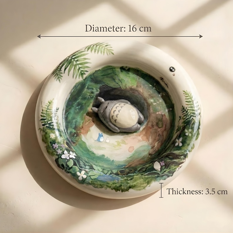 以下が含まれることがあります： 直径16cm、厚さ3.5cmの白い丸いセラミック皿。手描きの森の風景が描かれており、眠っているトトロ、シダ、花、小さな池が特徴です。