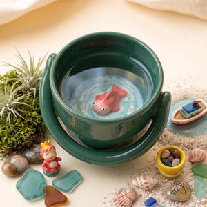 May include: A dark green ceramic bowl featuring a three-dimensional pink fish figurine at the bottom. The bowl is surrounded by decorative elements including a small character figurine, a miniature boat, seashells, sea glass, and moss.