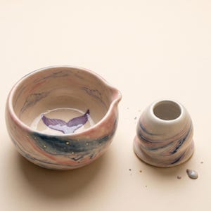 Puede incluir: Un cuenco de cerámica con pico vertedor y una taza pequeña a juego, ambos con un acabado jaspeado en rosa, azul y blanco con motas doradas. El interior del cuenco muestra una cola de ballena pintada en color púrpura en la base.