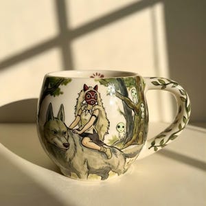 Könnte beinhalten: Keramikbecher mit handgemalter Illustration eines Charakters, der auf einem Wolf durch einen Wald reitet. Der Becher hat eine weiße Basis mit grünen und braunen Akzenten und einen mit grünen Blättern verzierten Griff.