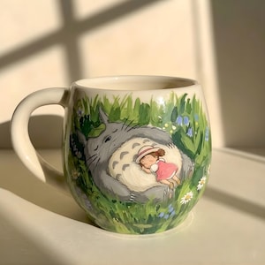 May include: A cream-colored ceramic mug featuring a hand-painted illustration of a large, grey, furry creature with a white belly sleeping in a field of green grass and small white and blue flowers, with a small child in a pink dress resting on it.