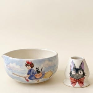 Może przedstawiać: Ceramiczna miska do mieszania i pasujący stojak na trzepaczkę z akwarelową ilustracją młodej osoby z czerwoną kokardą lecącej na miotle z czarnym kotem oraz stojak ozdobiony wizerunkiem pyszczka czarnego kota.