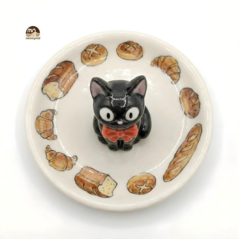 May include: A ceramic plate featuring a black cat figurine with a red bow tie, surrounded by illustrations of various baked goods. The plate has a white background and is perfect for serving small snacks or as a decorative piece.