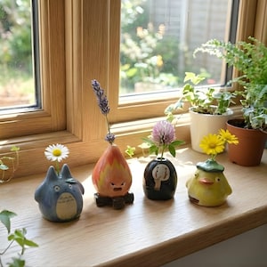 May include: A collection of four ceramic bud vases, each uniquely designed and holding a single flower or sprig. The vases are arranged on a wooden windowsill, with a Totoro-inspired vase, a flame-shaped vase, a black vase with a face, and a yellow duck-shaped vase.