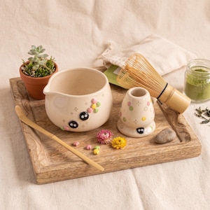 May include: A ceramic matcha bowl and whisk holder featuring small black soot sprite characters and colorful flower accents. The set includes a bamboo whisk and a bamboo scoop. A small succulent in a terracotta pot sits nearby.