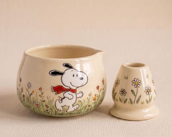 May include: A cream-colored ceramic creamer and a matching small candle holder. The creamer features a graphic of Snoopy wearing a red scarf, surrounded by painted green grass and small flowers. The candle holder has a matching floral pattern.