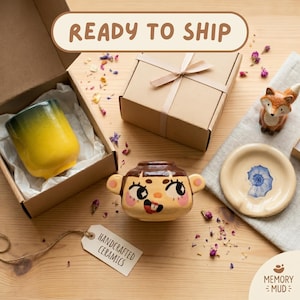 Puede incluir: Una taza de cerámica con degradado amarillo y verde en una caja de cartón abierta, un cuenco de cerámica marrón con cara de dibujos animados, un plato de cerámica beige con una ilustración de concha azul y una figura de zorro, con etiqueta HANDCRAFTED CERAMICS.