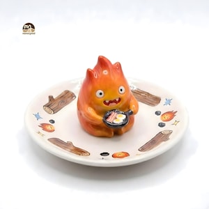 May include: A ceramic figurine of a fiery character holding a frying pan with bacon and eggs, set on a decorative plate. The plate features illustrations of logs, flames, and stars. The figurine is orange and yellow.