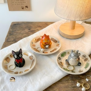 Könnte beinhalten: Drei kleine Keramik-Ringschalen mit Zeichentrickfiguren. Eine Schale zeigt eine schwarze Katze mit roter Schleife, eine andere einen Feuerdämon und die dritte einen grauen Waldgeist. Jede Schale ist mit Illustrationen zum Thema Essen und Natur verziert.