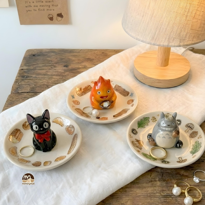 May include: Three small ceramic ring dishes featuring cartoon characters. One dish has a black cat with a red bow, another has a fire demon, and the third has a gray forest spirit. Each dish is decorated with food and nature-themed illustrations.
