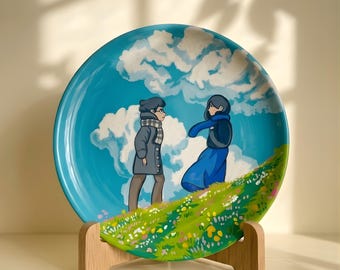May include: A round ceramic plate featuring a painted scene of two figures standing on a grassy hill against a bright blue sky with white clouds. One figure wears a gray coat and scarf, while the other wears a dark jacket and a long blue skirt.