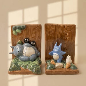 May include: A pair of ceramic bookends featuring a wood-grain texture. The left piece shows a large grey creature sleeping on green moss with two small black soot sprites. The right piece depicts a blue creature and a small white figure with acorns.