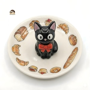 May include: A ceramic plate featuring a black cat figurine with a red bow tie, surrounded by illustrations of baked goods. The plate has a white background and the cat has large, round eyes. The plate is decorated with various types of bread and pastries.