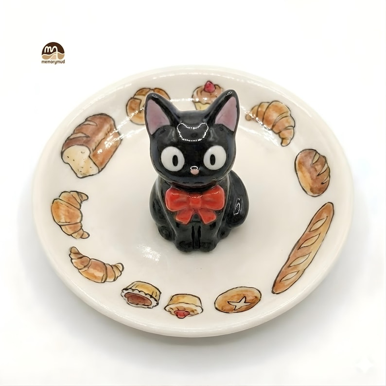 May include: A ceramic plate featuring a black cat figurine with a red bow tie, surrounded by illustrations of baked goods. The plate has a white background and the cat has large, round eyes. The plate is decorated with various types of bread and pastries.
