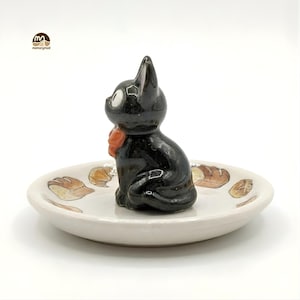 May include: A black cat figurine with a red bow sits on a small, off-white ceramic dish. The dish is decorated with brown and tan bread illustrations. The cat has white eyes and is in a sitting position. The dish is round.