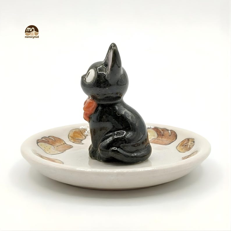 May include: A black cat figurine with a red bow sits on a small, off-white ceramic dish. The dish is decorated with brown and tan bread illustrations. The cat has white eyes and is in a sitting position. The dish is round.