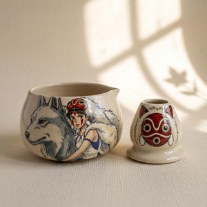 May include: A cream-colored ceramic bowl and a matching smaller ceramic stand. The bowl features a hand-painted illustration of a person with a red mask and a large grey wolf. The smaller stand displays a red and white mask design with pointed ears.