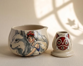 May include: A cream-colored ceramic bowl and a matching whisk holder. The bowl features a hand-painted illustration of a person with a red mask and a large grey wolf. The smaller whisk holder displays a detailed red and white mask design.