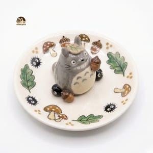 May include: A ceramic dish featuring a gray Totoro figure holding an acorn, surrounded by black soot sprites, mushrooms, and leaves. The dish is white with hand-painted details, perfect for a decorative display.