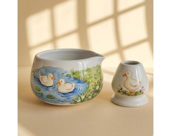 May include: A white ceramic creamer pitcher and a matching whisk holder, both decorated with hand-painted illustrations of yellow ducks. The pitcher features two ducks swimming in a blue pond, while the whisk holder shows a single standing duck.