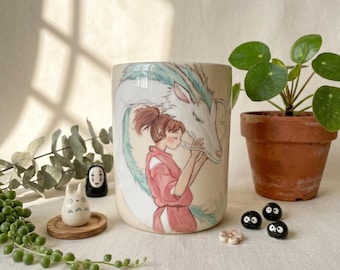 May include: A cylindrical ceramic mug featuring a colorful illustration of a person with brown hair in a pink garment embracing a long, white, and light blue dragon. The mug is surrounded by small decorative figurines and a green plant in a pot.