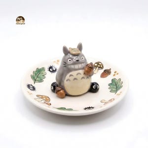 May include: A ceramic dish featuring a gray Totoro figure holding an acorn. The dish is white with painted green leaves, mushrooms, and other small characters. A charming decorative piece.