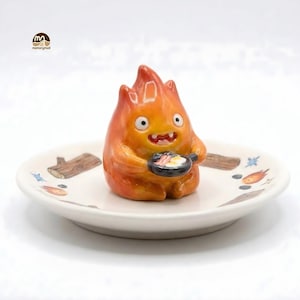 May include: A ceramic figurine of a fiery creature holding a frying pan with eggs and bacon, set on a decorative plate. The plate features illustrations of logs and flames. The figurine is orange and yellow with a smiling expression.