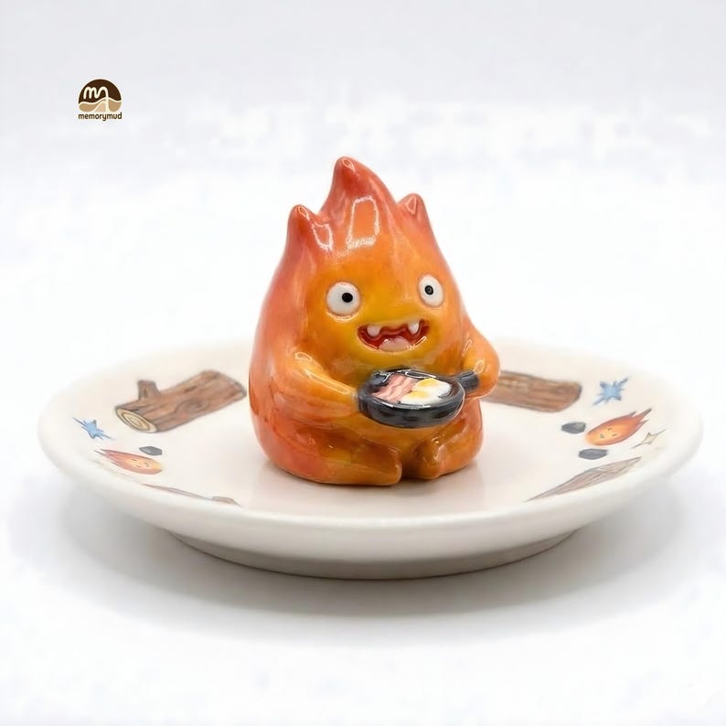 May include: A ceramic figurine of a fiery creature holding a frying pan with eggs and bacon, set on a decorative plate. The plate features illustrations of logs and flames. The figurine is orange and yellow with a smiling expression.
