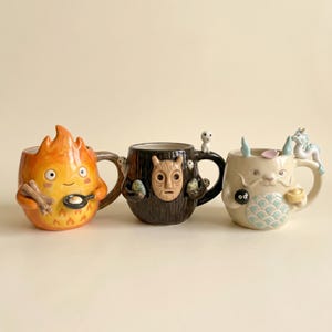 May include: Three ceramic mugs with unique character designs. One mug is shaped like a flame with a smiling face, another resembles a tree trunk with a mask, and the third has a dragon-like creature design. Each mug has a handle.