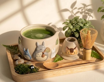 May include: A ceramic matcha bowl featuring embossed Totoro characters filled with green tea, a matching cat-themed whisk holder, a bamboo whisk, and a wooden spoon on a rectangular wooden tray with small decorative moss and stones.