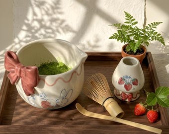 May include: A ceramic matcha bowl with a pink bow handle and rabbit illustration, a bamboo whisk, a bamboo scoop, and a ceramic whisk holder decorated with strawberries. The bowl contains green matcha powder, and a small fern plant is nearby.