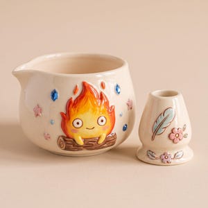 Puede incluir: Un cuenco de cerámica color crema con pico vertedor, que presenta un personaje de llama naranja y amarillo en relieve sobre un tronco, acentuado con pequeñas estrellas rosas y gemas azules. A su lado hay un soporte para batidor a juego.