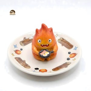 May include: A ceramic figurine of a fiery character holding a frying pan with eggs and bacon, set on a decorative plate. The plate features illustrations of logs, flames, and stars. The figurine is orange with a smiling face.