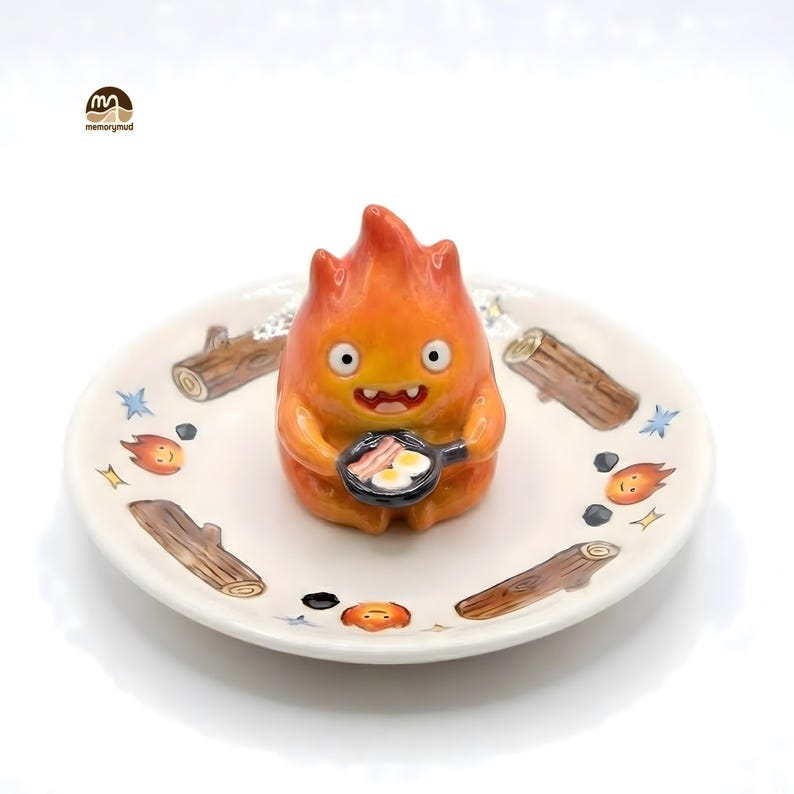 May include: A ceramic figurine of a fiery character holding a frying pan with eggs and bacon, set on a decorative plate. The plate features illustrations of logs, flames, and stars. The figurine is orange with a smiling face.