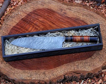 May include: A Damascus steel chef's knife with a patterned blade, presented in a black wooden box with decorative trim. The handle features a combination of dark and light wood tones. The knife rests on a bed of straw.