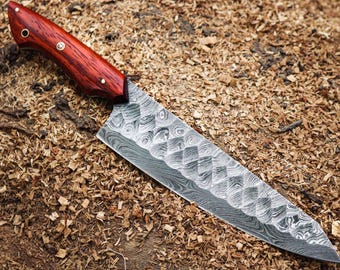 May include: A Damascus steel chef's knife with a patterned blade and a reddish-brown wooden handle. The knife is lying on a bed of wood shavings, showcasing its craftsmanship and design.
