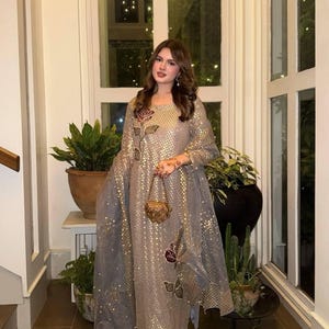 Puede incluir: Conjunto formal con una túnica dorada brillante y pantalones a juego. La túnica está adornada con bordados florales y se combina con una bufanda gris transparente. Un pequeño bolso dorado completa el look.