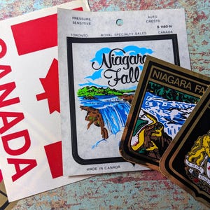 May include: A collection of vintage Canadian stickers, including a red and white Canadian flag, a sticker with the text "CANADA" in red, a sticker with the text "Niagara Falls" and a picture of the falls, and a sticker with the Canadian coat of arms.