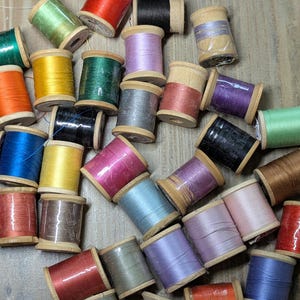 May include: A collection of colorful thread spools in various shades, including red, orange, green, blue, and purple. The spools are made of wood and filled with thread, perfect for sewing and crafting projects.