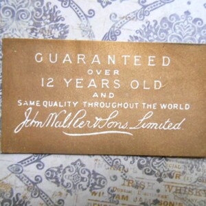 May include: A brown paper label with white text that reads "GUARANTEED OVER 12 YEARS OLD AND SAME QUALITY THROUGHOUT THE WORLD John Walker Sons Limited."