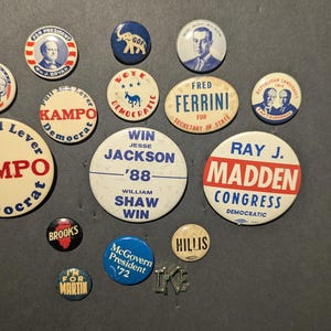 May include: A collection of vintage political campaign buttons, featuring various candidates and slogans. The buttons are round and oval, with text and images in red, white, blue, and black. Some buttons include the names "KAMPO", "MADDEN", and "JACKSON".