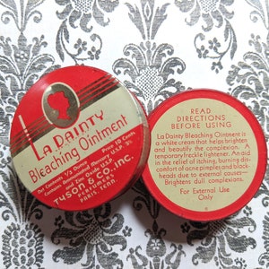 May include: Two red and white metal tins of La Dainty Bleaching Ointment. The front tin shows a woman's silhouette and the text "La Dainty Bleaching Ointment". The back tin has directions for use, including "La Dainty Bleaching Ointment is a white cream that helps brighten and beautify the complexion."