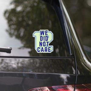 May include: A rectangular sticker with the text "WE DID NOT CARE" in a blue and white font, with a light green number 12. The sticker is affixed to the rear window of a black car.