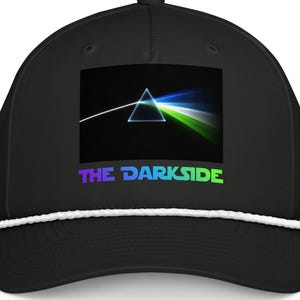 Puede incluir: Gorra de béisbol negra con un gráfico de un prisma que refracta la luz y un detalle de cuerda blanca en el borde. El texto "THE DARKSIDE" se muestra en un degradado de colores. Un accesorio de moda.