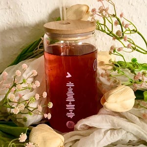 Könnte beinhalten: Ein klares Glas mit einem Holzdeckel und einem durchsichtigen Strohhalm, gefüllt mit einer roten Flüssigkeit. Das Glas hat ein weißes grafisches Design. Das Glas ist von weißen Blumen und Grün umgeben.